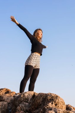 Bir kadın dışarıda duruyor, yoga pozu vererek kollarını gökyüzüne doğru açıyor. Ayar parlak mavi bir gökyüzü ve güneş ışıldıyor. Çalışması için sakin bir atmosfer yaratıyor..