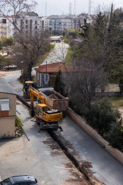 Türkiye, Antalya - 10.03.2025: Sakin bir banliyö mahallesinde dar bir sokakta ağır makineler çalışıyor. İnşaat ekipleri kazı yaparken, sarı kazıcı enkazı kamyona yüklüyor.