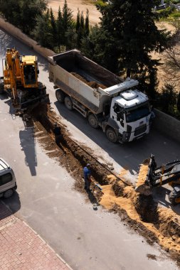 Türkiye, Antalya - 10.03.2025: Kaldırım boyunca ağır makineler çukur kazıyor. kepçe doldurucu aktif olarak yol onarımı için hazırlanıyor. Yakınlarda araçlar park halinde, aktiviteyi izliyorlar..