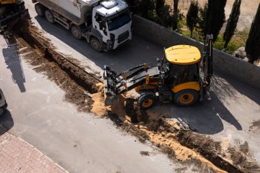 İnşaat işçileri büyük bir kazıcı kullanarak yol kenarında bir çukur kazıyorlar. Kamyonlar yakınlarda park halinde ve güneş gün boyunca olay yerini aydınlatıyor. Toz ve toprak görünür. Hava görünümü