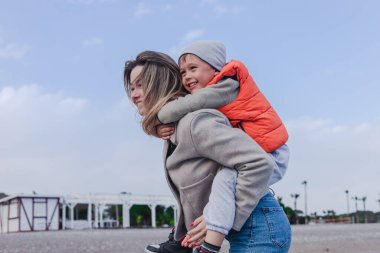 Anne, neşeli oğlunu kışın kumsalda sırtında taşıyor. Gökyüzü bulutlu ve gülümsemeleri kıyı şeridini keşfederken paylaştıkları bağı vurguluyor. Annelik ve aktif aile zamanı