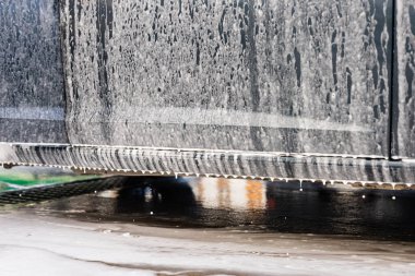 İyice yıkanan, köpükle kaplanmış bir araba. Köpükler kenarlardan taşar ve su beton yüzeye damlar. Parlak güneş ışığı temizlik sürecini aydınlatır. self-servis araba yıkama