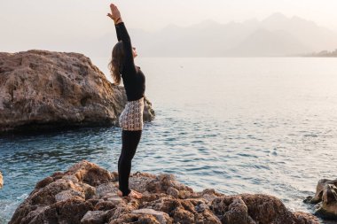 Bir kadın, güneş doğarken kayalık bir sahilde yoga pozu veriyor. Sakin sular şafağın yumuşak renklerini yansıtırken kayalıklar ihtişamlı bir şekilde arka planda duruyor ve sakin bir atmosfer yaratıyor..