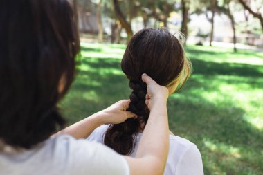 Bir kadın, ağaçlarla çevrili yemyeşil bir parkta otururken kızlarının saçını dikkatlice örüyor. Güneş ışığı yaprakların arasından süzülerek sakin bir atmosfer yaratır..