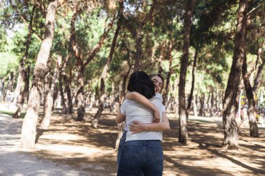 İki arkadaş, uzun ağaçlarla dolu güneşli bir orman parkında birbirlerine sevinçle sarılıyorlar. Arkadaşlıklarını gülümseme ve kahkahalarla kutlarken atmosfer sıcak ve canlı..
