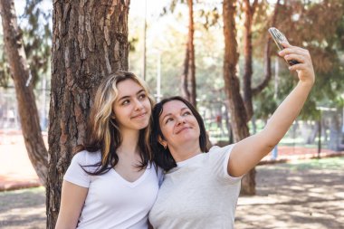 Ağaçlarla dolu canlı bir parkta, iki kadın, diğer ve genç kız selfie için birlikte poz veriyorlar. Biri uzun, dalgalı saçlara sahipken, diğeri parlak bir şekilde gülümsüyor, ikisi de güneşli bir günün keyfini çıkarıyor.