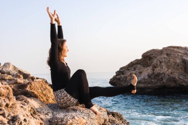 Gülümseyen kadın deniz kenarındaki kayaya oturmuş, kolları havada yoga pozu veriyor ve eğleniyor. Sakin su ve yumuşak gün batımı etrafında huzurlu ve mutlu bir atmosfer yaratır..