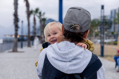 Yetişkin bir büyükannenin sırtında taşınırken sarı saçlı neşeli çocuk gülümsüyor. Palmiye ağaçları ve bulutlu gökyüzünün altındaki uzak dağlarla dolu manzaralı bir gezinti alanı boyunca yürüyorlar..