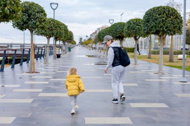 Ebeveyn büyükanne, ağaçlarla kaplı geniş bir gezinti alanında küçük bir çocukla el ele yürüyor. Çocuk, sarı ceketli, bulutlu bir günde sevdikleriyle anın tadını çıkartıyor..