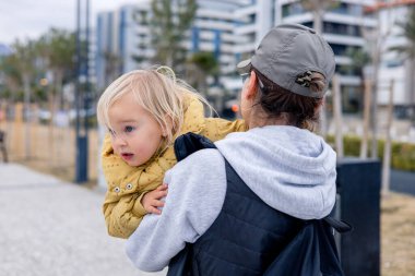 Yetişkin bir büyükannenin sırtında taşınırken sarı saçlı neşeli çocuk gülümsüyor. Palmiye ağaçları ve bulutlu gökyüzünün altındaki uzak dağlarla dolu manzaralı bir gezinti alanı boyunca yürüyorlar..