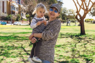 Sunny Park 'ta elinde küçük bir kız torunu olan ve ona meraklı bir şekilde bakan bir kadın var. Çevrede yeşil çimenler ve ağaçlar yer alır ve arka planda binalar görülebilir..