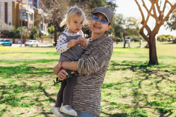 Sunny Park 'ta elinde küçük bir kız torunu olan ve ona meraklı bir şekilde bakan bir kadın var. Çevrede yeşil çimenler ve ağaçlar yer alır ve arka planda binalar görülebilir..