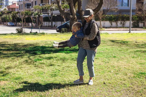 Kadın büyükannesi, genç kızıyla canlı yeşil parkta eğlenmekten zevk alıyor. Güneş, öğleden sonraları ağaçlar ve açık alanla çevrili bir şekilde gülüşlerini paylaşırken ışıl ışıl parlıyor..