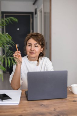 Mutlu gülümseyen olgun yatırımcı kadının portresi beyaz elbiseli, karı kutlayan, emeklilik tasarrufu yapan, kazanan el bilgisayarda oturan, masada yeşil çiçekli.