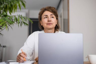 Beyaz elbiseli, düşünceli, olgun bir iş kadınının portresi. Düşünme ya da karar verme, dizüstü bilgisayar kullanma, elinde kalem tutma, yaratıcı fikirler üzerinde beyin fırtınası, problem çözme. 