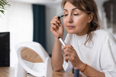 Orta yaşlı bir kadın, parlak, rahat bir odada, ahşap bir masada otururken maskara sürerek güne hazırlanıyor. Ayna, güzelliğini arttırırken odaklanmış yüz ifadesini yansıtıyor. Sabah rutini