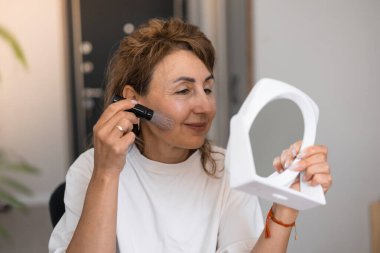 Orta boylu, olgun, güzel bir kadın evde oturan bir aynaya bakarken fırça kullanarak makyaj yapıyor. Gerçek insanlar ve günlük rutin konsept