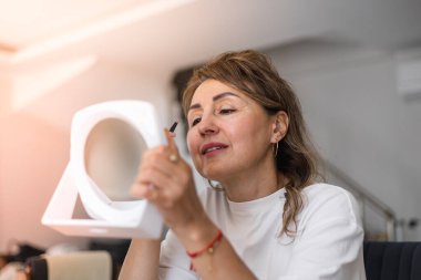 Bir kadın şık bir odada oturuyor, aynayla, cımbızla ve kaş aldırarak. Doğal ışık, makyaj rutinini hassasiyetle arttırırken odaklanmış yüz ifadesini aydınlatır..