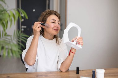 Orta boylu, olgun, güzel bir kadın evde oturan bir aynaya bakarken fırça kullanarak makyaj yapıyor. Gerçek insanlar ve günlük rutin konsept