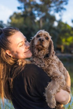 Bir kadının cocker spaniel 'ını göğsüne yasladığı neşeli bir an. Bağları yemyeşil ve güneşli bir gökyüzünün altında, bağlarını yansıtan sakin bir göl arasında parlıyor..