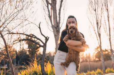 Bir kadın cocker spaniel köpeğini sevgiyle tutarken parkta huzurlu bir günbatımı yürüyüşünün tadını çıkarıyor. Sahne, aralarındaki bağı güzel bir doğal zemin ile yakalıyor..