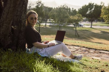 Parktaki bir ağaca yaslanmış bir kadın cocker spaniel köpeği yakınlarda dinlenirken dizüstü bilgisayarını kullanıyor. Güneş parlıyor ve çevre yeşilliklerle dolup taşıyor. Her ikisi de bir anı paylaşıyor..