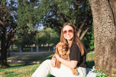 Güneşli bir parkta çimlerin üzerinde oturan bir kadın cocker spaniel 'ini sevgiyle kucaklıyor. Her ikisi de ağaçlar ve doğa ile çevrili sakin ve sakin ortamlarında rahat ve mutlu görünüyorlar..