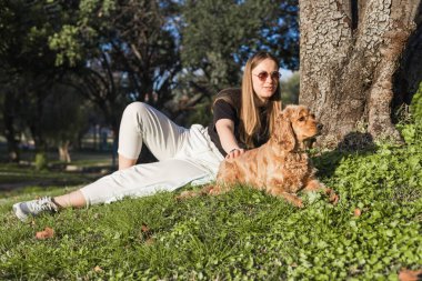 Bir kadın parktaki çimlerin üzerinde rahatlar, cocker spaniel cinsiyle sevgiyle etkileşim içindedir. Güneşli bir öğleden sonra bir ağacın gölgesinde huzurlu bir an yaşarlar..