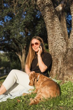 Kadın ve evcil hayvan parkta bir ağacın altında oturuyor, gündelik giyinmiş ve güneş gözlüğü takıyor, telefonda konuşurken. Cocker köpeği çimlerin üzerinde dinleniyor, güneşli öğleden sonranın tadını çıkarıyor..