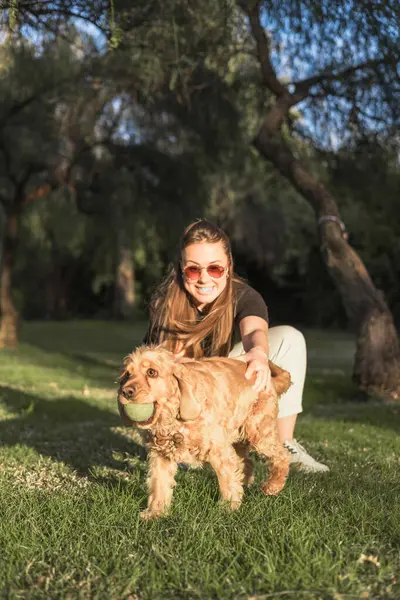 Çimlere çömelmiş bir kadın cocker spaniel köpeği ile etkileşime girerken geniş bir gülümseme sergiliyor. Sahne, yemyeşil bir park ortamında ikisinin arasında bir neşe ve bağlantı anı yakalıyor. Hayvan eğitimi süreci