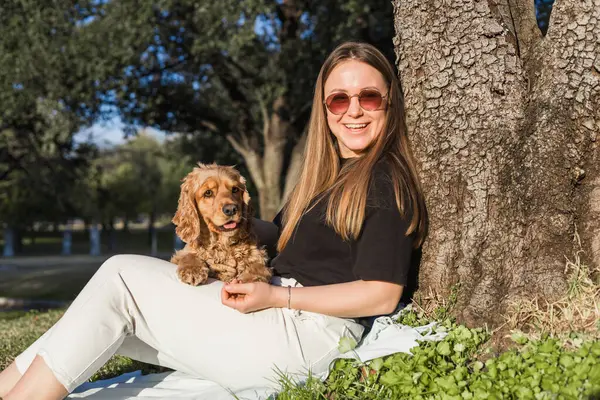 Bir kadın parktaki çimlerin üzerinde rahatlar, cocker spaniel cinsiyle sevgiyle etkileşim içindedir. Güneşli bir öğleden sonra bir ağacın gölgesinde huzurlu bir an yaşarlar..