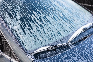 Köpük ve köpük birikerken, parlak güneş ışığı sabunlu bir arabanın ön camından yansıyor. Garaj yolu huzurlu bir atmosferle çevrilidir. Bu da öğleden sonra yıkanma etkinliğinin göstergesidir..