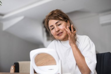 Bir kadın rahatlamış bir iç mekanda masada otururken yüzüne dikkatlice nemlendirici sürüyor. Doğal ışık odayı dolduruyor, odaklanmış ifadesini ve kişisel bakım rutinini vurguluyor..