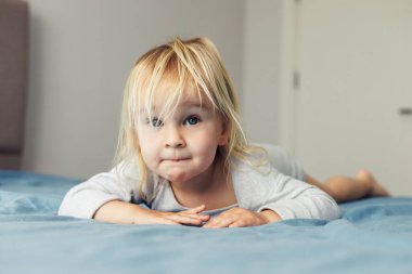 Neşeli sarışın bir çocuk evdeki bir yatakta neşeyle oynuyor. Büyük bir gülümsemeyle öne eğiliyor, neşeli ruhunu gün boyunca rahat ve rahat bir ortamda sergiliyor..