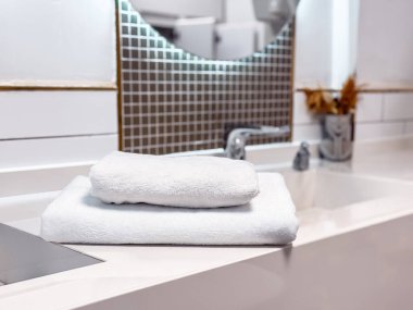 Çağdaş bir banyoda temiz beyaz havlular şık bir tezgahın üzerinde durur. Minimalist tasarım, aydınlık ve temiz atmosferi tamamlayarak, bir sakinlik ve hijyen duygusunu vurguluyor..