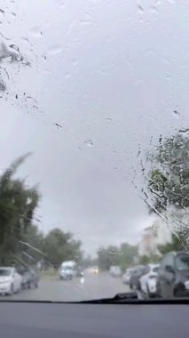 Yağmur fırtınası sırasında şehirde araba kullanmak. Yağmurda araba sürerken arabanın ön camına yağmur damlaları düşer. Bir arabanın içinden görüntüyü al. Araba silecekleri çabuk çalışır.