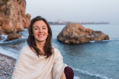 Genç, beyaz, gülümseyen kadın, yumuşak dalgalarla çakıllı sahilde oturuyor. Güneş uzak şehrin ardından batarken düşünceli bir şekilde ufka bakıyor ve sakin bir atmosfer yaratıyor. Tek başına seyahat kavramı