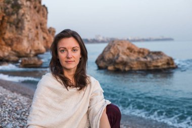 Genç, gülümseyen bir kadın, ayaklarında hafif dalgalarla çakıllı bir plajda oturuyor. Güneş uzak şehrin ardından batarken düşünceli bir şekilde ufka bakıyor ve sakin bir atmosfer yaratıyor..