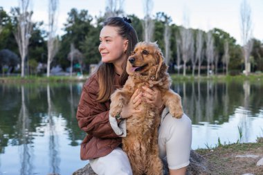 Kadın, sakin bir göletin yanında çömelirken nazikçe cocker spaniel 'ini okşar. Hayvan dili dışarıda, huzurlu bir park ortamında sıcak güneşin tadını çıkarırken mutlu görünüyor. Kucaklayan ve sarılan köpek