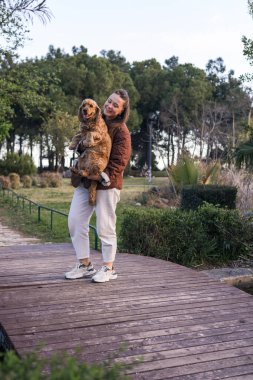 Bir kadın parktaki ahşap bir patikada dikilirken bir cocker spaniel 'i yakından tutar. Ağaçlar ve yeşilliklerle çevrili olarak, birlikte sıcak bir günün tadını çıkarırlar, bağlarını vurgularlar..