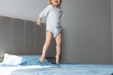 Sarışın küçük bir kız evde oyun oynamaktan zevk alıyor, enerjik bir şekilde yatakta zıplıyor. Yumuşak öğleden sonra ışığı sıcak bir atmosfer yaratıyor. Sıcak yatak odasında, neşeli aktivitelerini vurguluyor.. 