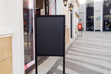 Bir alışveriş bölgesindeki kaldırımda boş bir tabela duruyor. Çevrede modern mimari ve mağazalar yer alıyor. Parlak gün ışığı canlı bir atmosfer yaratır..