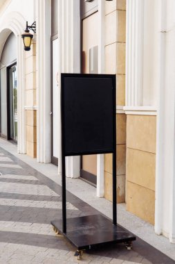 Mockup, siyah kaldırım levhası hareketli bir şehir caddesinde. Etraftaki dükkanlar ve palmiye ağaçları, yoldan geçenler için davetkar bir ortam yaratıyor..