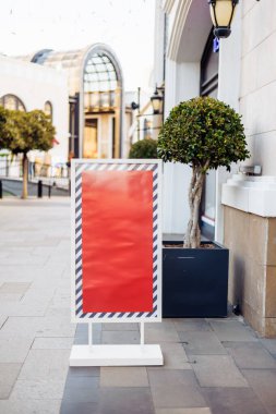 Arka planda düzgün budanmış ağaçlar ve çağdaş binalarla çevrili boş reklam panosu kaldırıma yerleştiriliyor. Tahta gün ışığında açıkça görülüyor, dikkat çekiyor..