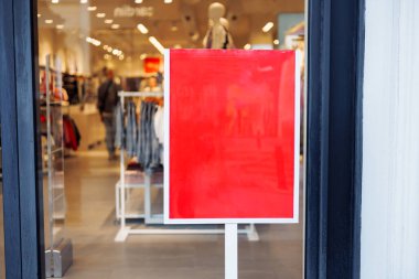 Dolu bir alışveriş bölgesindeki bir giyim mağazasının önünde duran parlak kırmızı tabela, mağazanın geçici olarak kapalı olduğunun sinyalini veriyor. İçerisi giysi ve manken vitrinleriyle dolu..