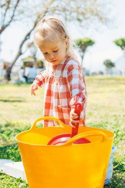İşaretli elbiseli küçük bir çocuk parktaki çimenli bir arazide büyük bir turuncu kovanın içine bakmak için eğilir. Açık hava yaz aktiviteleri ve su durin pikniğiyle eğlenceli oyunlar