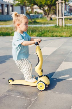 At kuyruklu genç bir çocuk parktaki kaldırımlı bir yolda sarı bir scooter kullanıyor. Çevreleyen yeşillik ve oyun alanı ekipmanları bu aydınlık günde neşeli bir atmosfer yaratır..