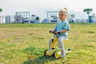 Küçük bir çocuk açık gökyüzünün altındaki çimenli bir parkta parlak sarı bir scooter sürmekten hoşlanıyor. Arkaplanda ağaçlar ve binalar var, neşeli bir atmosfer yaratıyorlar..