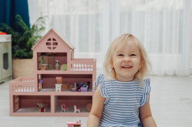 Açık renk saçlı küçük bir çocuk, canlı oyuncak evin yanındaki rahat mekanda oyun oynamaktan zevk alıyor..