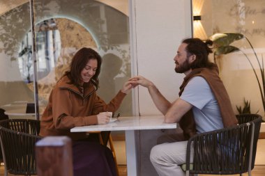 Bir çift şık bir kafede karşılıklı oturuyor, gülümsüyor ve sıcak bir sohbete girişiyor. Atmosfer, kapalı yeşil alan ve yumuşak ışıklandırma ile rahatladı. Ellerini tutuyorlar.